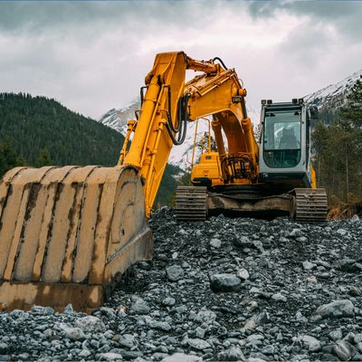 Yellow excavator on rocky terrain with forest and mountains in the background.