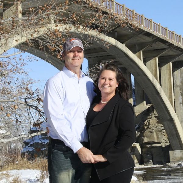 A smiling couple holding hands under a concrete arch bridge on a sunny winter day.