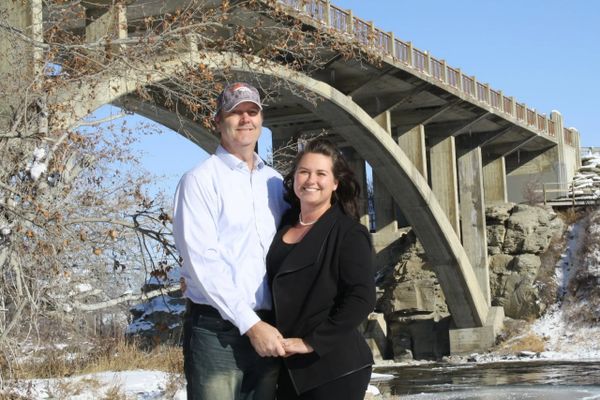 A smiling couple holding hands under a concrete arch bridge on a sunny winter day.