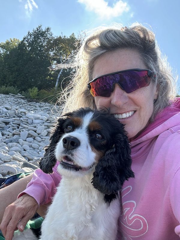 Woman in pink hoodie with sunglasses poses happily with a black and white dog on a rocky beach.