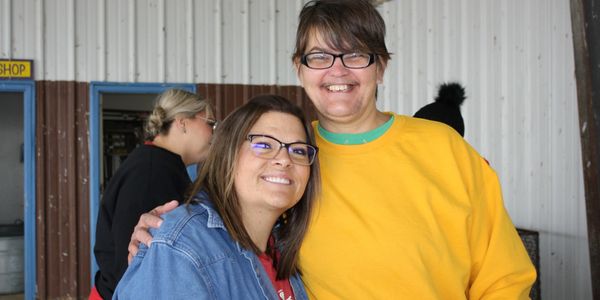 Two smiling women posing together, one in a yellow sweatshirt and the other in a denim jacket.