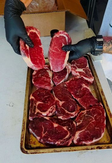 Two hands in black gloves holding raw steaks over a tray of steaks.