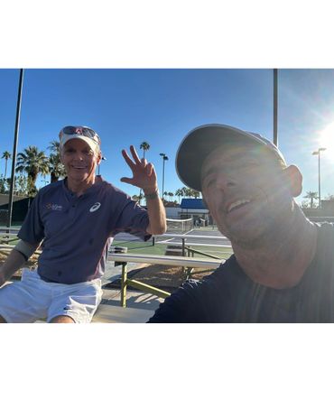 Two men smiling and posing at a sunny outdoor tennis court.