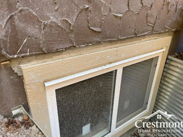 Close-up of a basement window with damaged stucco and dirt buildup.