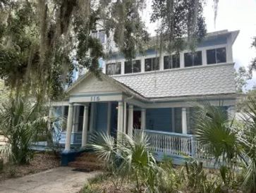 Blue house with large front porch surrounded by greenery.