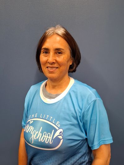 Smiling woman in a blue 'The Little Gym School' t-shirt stands against a gray wall.
