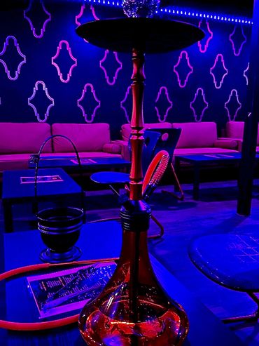 A red hookah on a table in a neon-lit lounge with purple sofas and patterned walls.