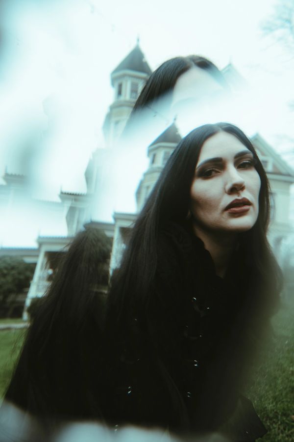 A woman with long dark hair stands in front of a vintage building, with a dreamy double exposure effect.