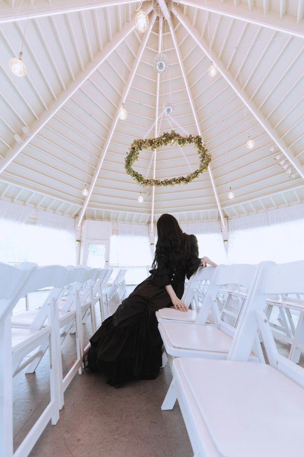 A woman in a black dress sits alone among white chairs in a bright, elegant venue.