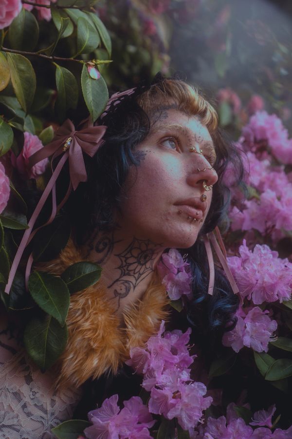 Person with tattoos and piercings surrounded by pink flowers and green leaves.