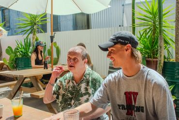 Two men enjoying a sunny outdoor cafe with drinks and plants around.