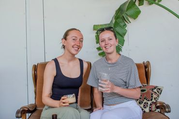 Two young women smiling and holding drinks while sitting on a couch indoors.