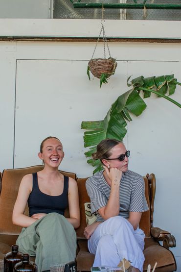 Two women sitting on a brown couch, one smiling and the other wearing sunglasses.