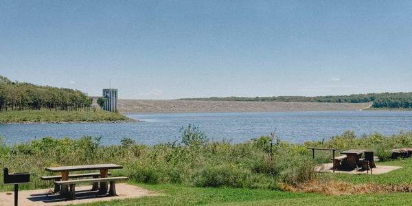 Picnic tables by a lake with a dam in the distance on a clear sunny day.