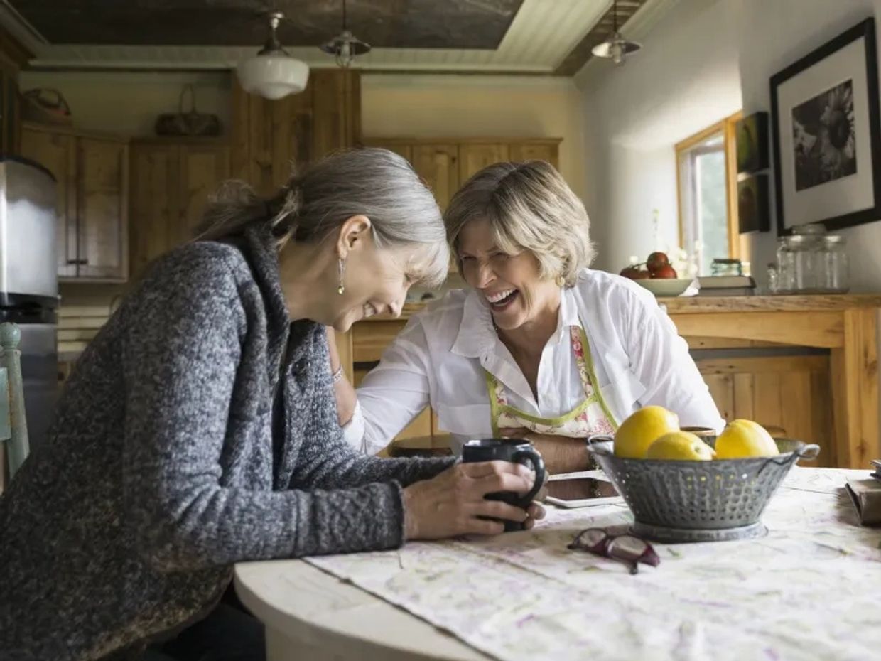 Two older women laughing together at a kitchen table.