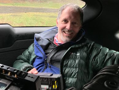 Smiling man in a green jacket sitting in a vehicle with camera equipment.