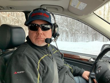 Man wearing sunglasses and headset inside a car in a snowy environment.