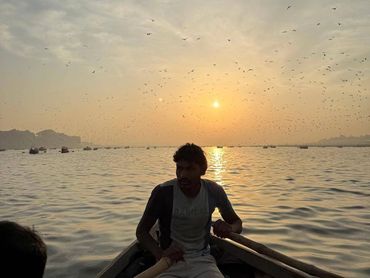 Man rowing a boat at sunset with birds flying overhead.