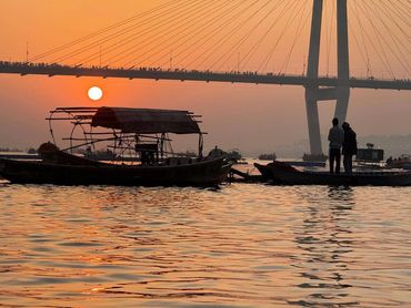 Silhouetted boats and people on a river at sunset under a suspension bridge.