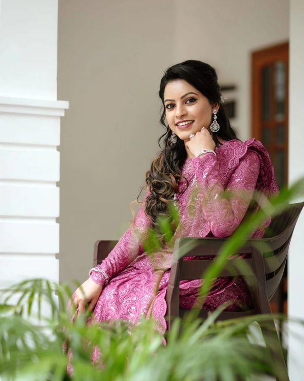 A smiling woman in a pink traditional dress sits gracefully on a chair indoors.
