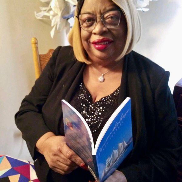 A smiling woman with blonde hair and glasses reads a book indoors.