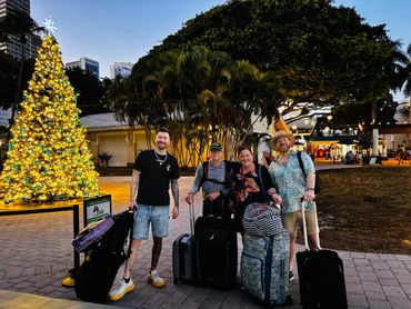Four people with luggage standing near a decorated Christmas tree in an outdoor setting at dusk.