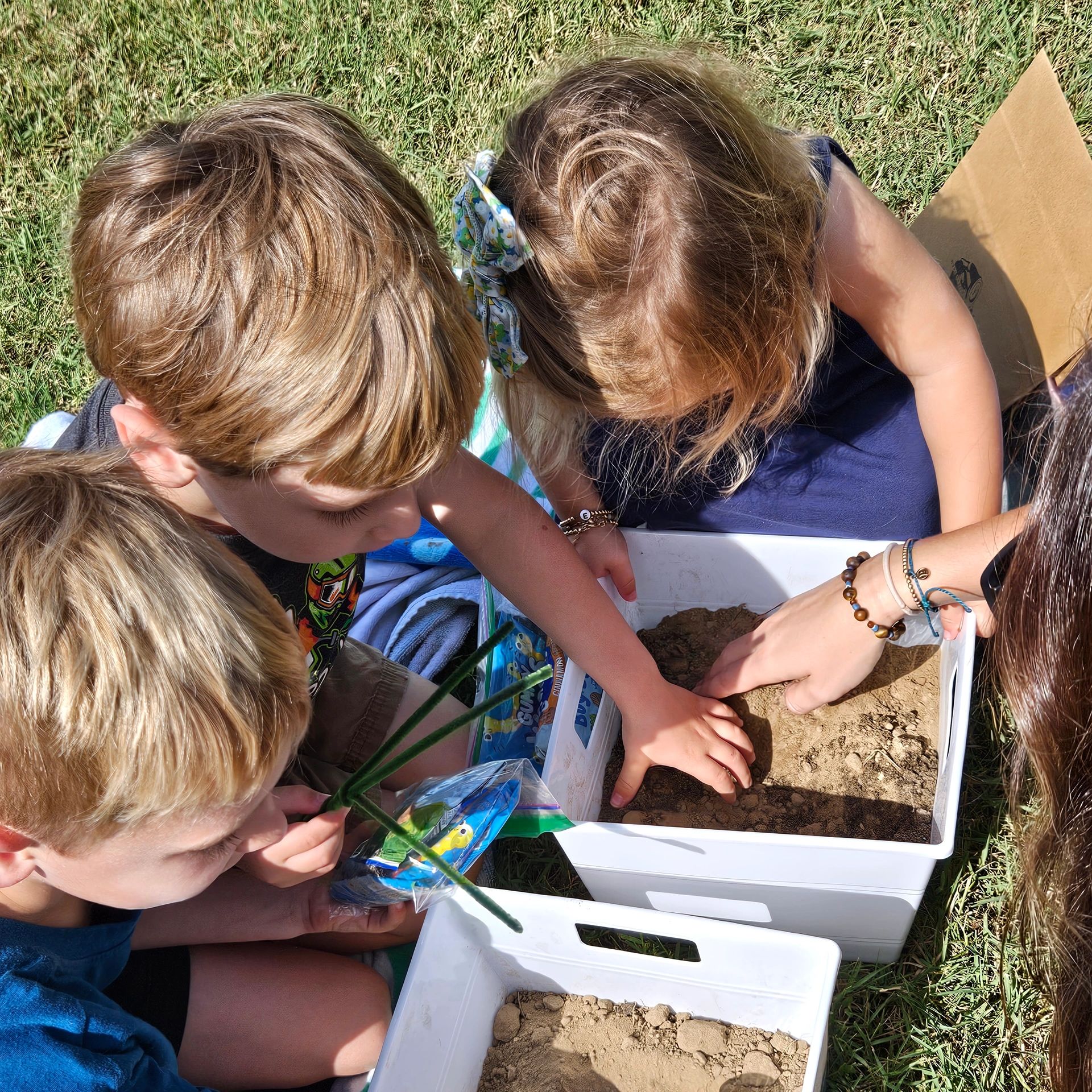Children exploring soil in white containers outdoors on grass.