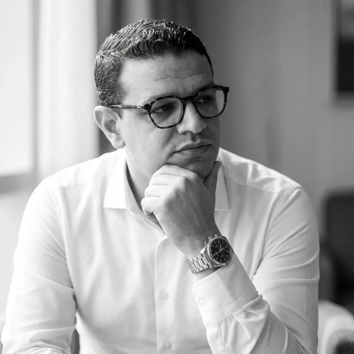 Yassine Sellami in glasses and white shirt looking thoughtful, resting chin on hand.
