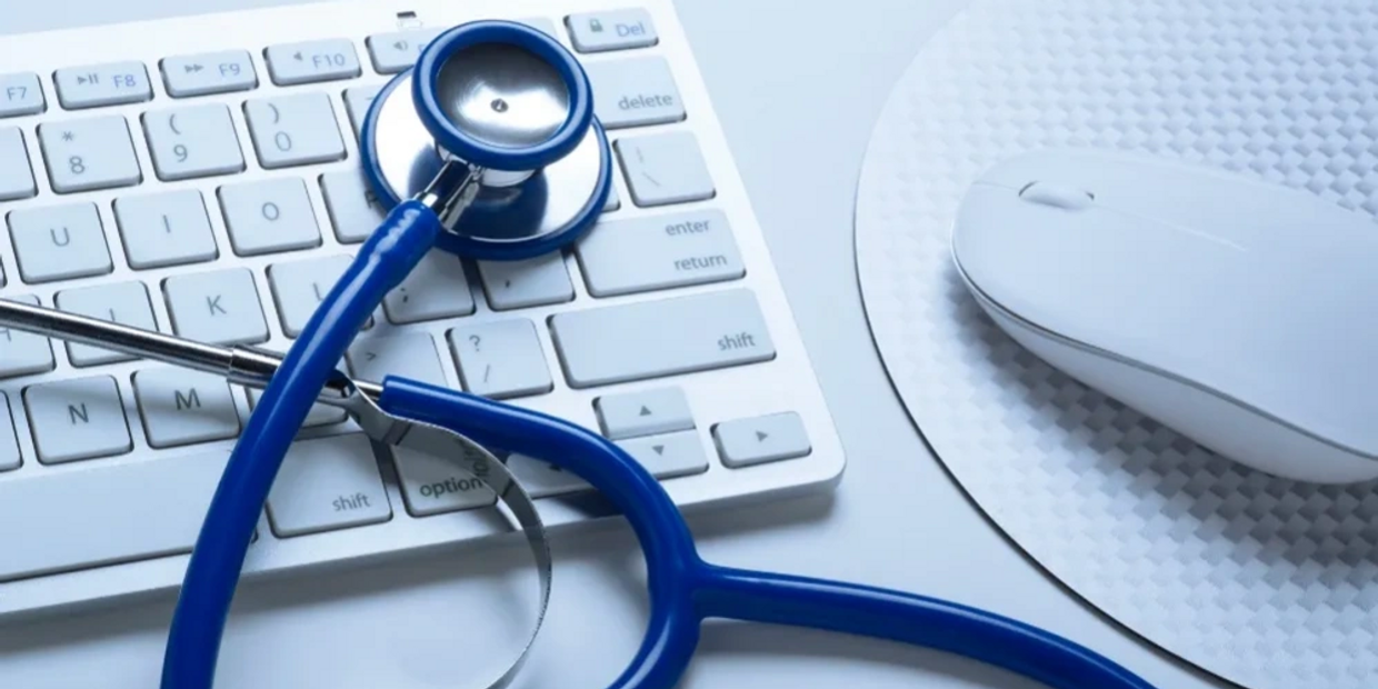 Blue stethoscope laying on top of white keyboard, white mouse and mouse pad on table.
