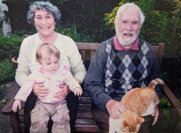Elderly couple with a toddler and a cat on a wooden bench outdoors.