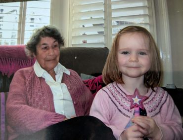 A young girl holding a star-shaped wand sits beside an elderly woman in a cozy living room.