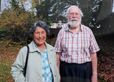 Smiling elderly couple standing outdoors in autumn with trees and house in background.