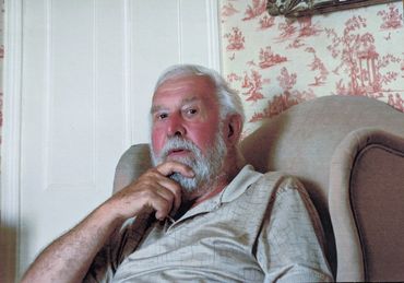Elderly man with white beard sitting thoughtfully in a beige armchair.