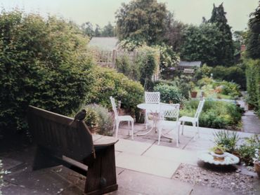 A peaceful garden patio with white chairs, a wooden bench, and a bird perched on it.