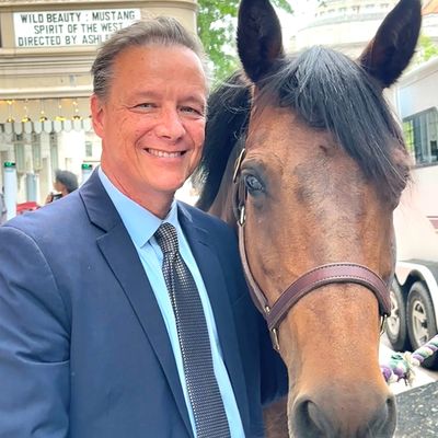 Man in suit smiling next to a brown horse outdoors.