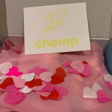 A card with candies and the word 'chomp' on pink tissue paper with heart confetti.