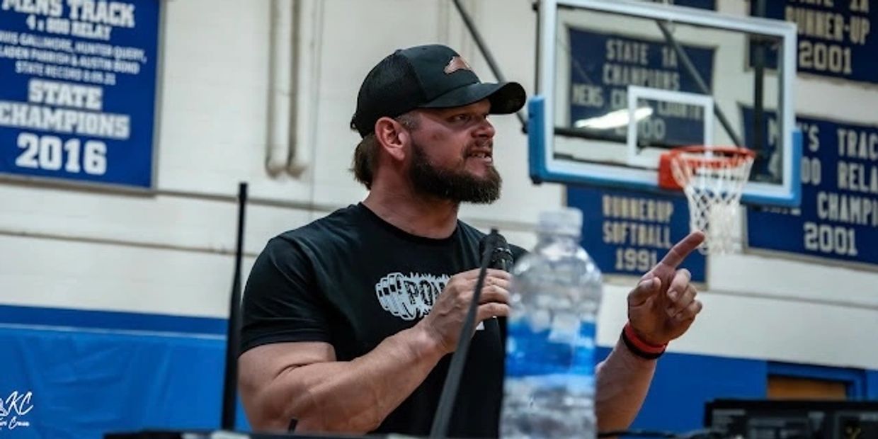 Man speaking passionately in a gymnasium with sports banners in the background.