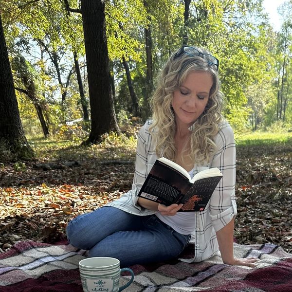 Woman reading a book outdoors on a blanket in a wooded area.