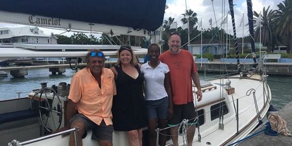 Camelot Segelboot am Dock, davor 4 lächelnde Personen,
2 Männer und 2 Frauen, im Hintergrund Palmen