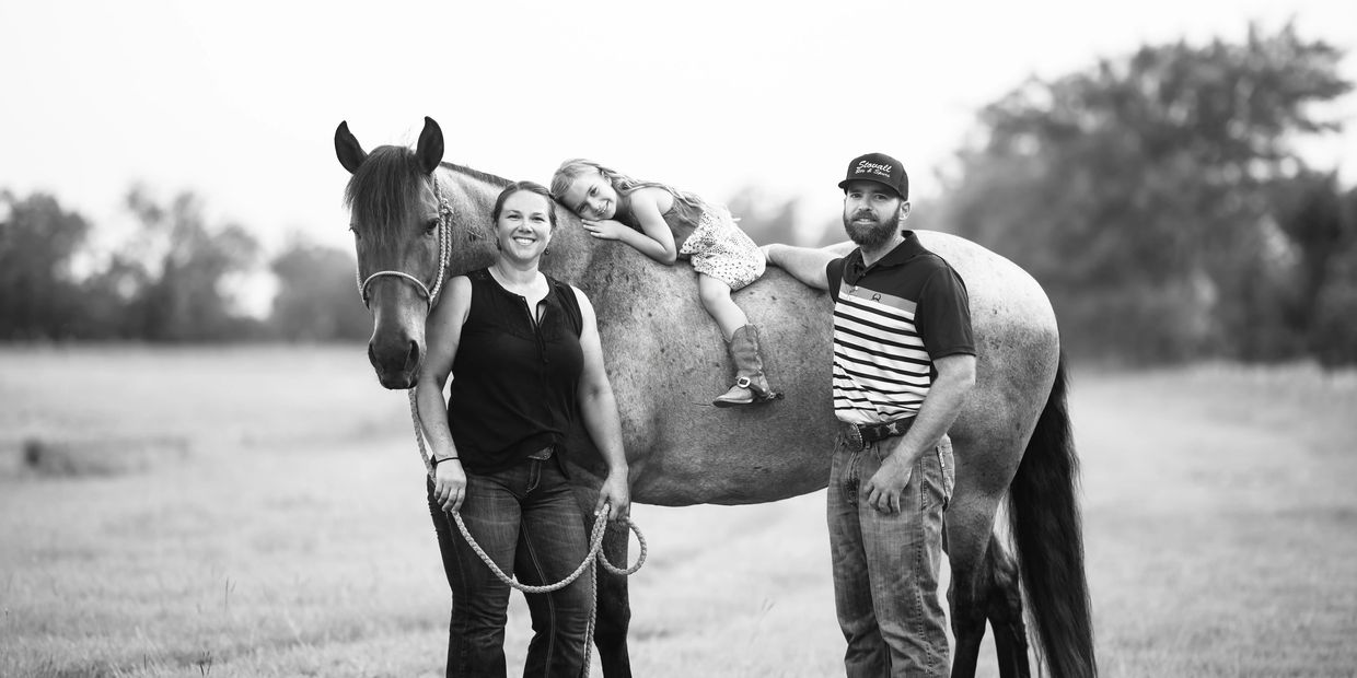 Family portrait with a horse in a field, featuring a smiling woman, a man, and a child lying on the horse.