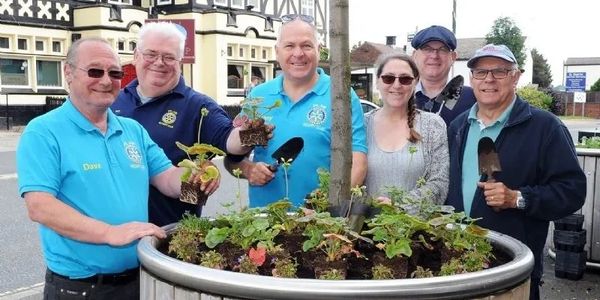 A group of six people planting flowers in a large wooden planter outdoors.