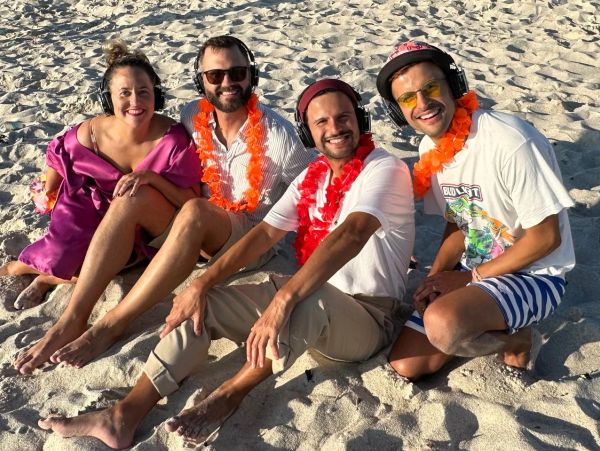Four friends with leis and headphones smiling on the beach sand.