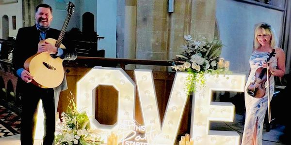 Guitarist and violinist standing by illuminated letters saying "Love"
