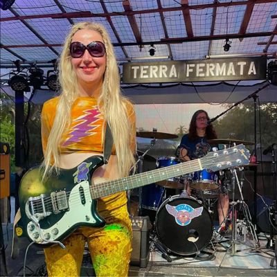 Blonde guitarist smiling on stage with drummer behind at Terra Fermata.