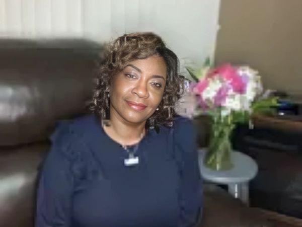 A woman in a dark outfit sits on a leather couch with a flower vase in the background.