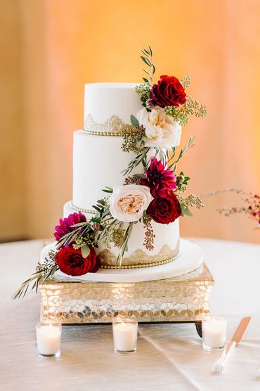 Three tier wedding cake and gold accents with red roses, burgundy dahlias and white garden roses