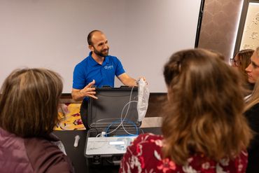 Man in blue shirt in back of briefcase that has products he is showing to attendees at workshop