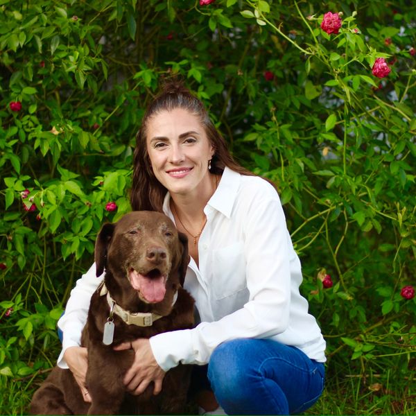 photo of woman kneeling down next to chocolate lab smiling in front of green and pink flowered bush
