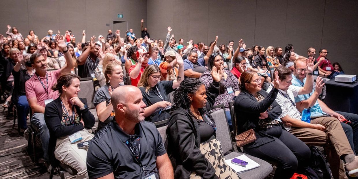 crowd at conference sit with some raising their hands. All look towards presetner (off screen)