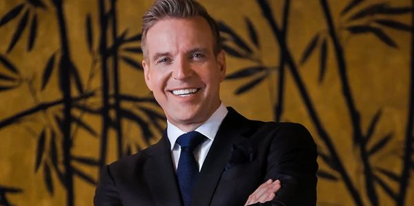 Smiling man in a suit with arms crossed against a patterned background.